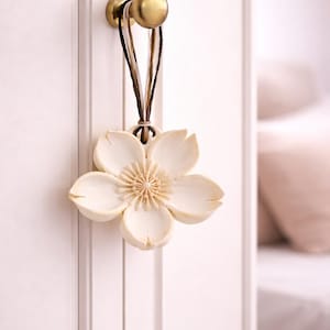 Könnte beinhalten: Cremefarbener, blütenförmiger dekorativer Hängeornament. Das Ornament wird von einer braunen und beigefarbenen Schnur an einem goldfarbenen Knauf an einer weißen Tür aufgehängt. Die Blume hat detaillierte Blütenblätter und ein strukturiertes Zentrum.