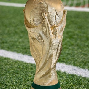 Pode incluir: Um troféu dourado da Copa do Mundo FIFA, apresentando um globo terrestre sustentado por duas figuras, repousa sobre uma base verde com a inscrição "FIFA WORLD CUP". O troféu está em um campo verde com linhas brancas.