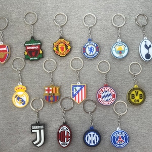 May include: Collection of keychains featuring various soccer team logos, including Arsenal, Liverpool, Chelsea, and Manchester City. Each keychain has a metal ring and a colorful, detailed emblem.