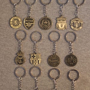 Op de afbeelding: Een verzameling metalen sleutelhangers met logo's van verschillende voetbalteams. De sleutelhangers hebben een zilveren ring en ketting, met logo's zoals Chelsea, Arsenal, Manchester United en Liverpool. Op een grijze ondergrond.