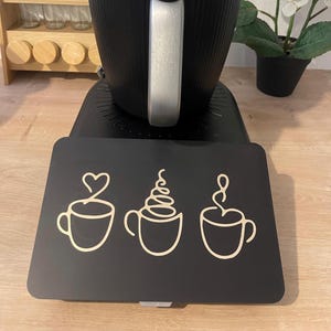 May include: A black coffee maker sits on a black mat with three coffee cup designs in a light beige color. The coffee cup designs include a heart, whipped cream, and steam. A wooden spice rack and a potted plant are in the background.