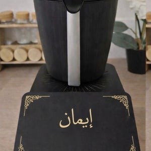May include: Black electric rice cooker with a silver handle, sitting on a black base. The base has the Arabic word "إيمان" in gold lettering. The cooker is in front of a shelf with jars and a plant.