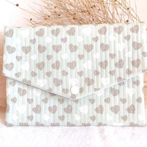 May include: A light blue, quilted fabric clutch with a flap closure. The clutch is patterned with white and taupe hearts. A white button secures the flap. The background includes a sprig of dried flowers and a sheer pink fabric.