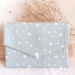 Pode incluir: Embreagem acolchoada azul claro com um padrão de estrelas brancas. A embreagem tem um fecho de aba com um botão branco. O fundo inclui plantas secas e tecido rosa.