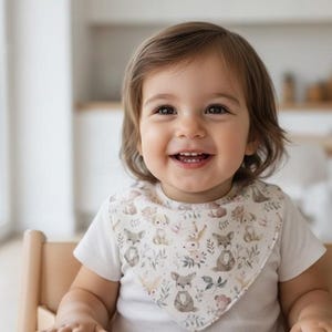 May include: A smiling toddler wearing a white t-shirt and a bib with a woodland animal pattern. The child is seated in a wooden high chair, with a bright kitchen in the background. The bib features foxes, bears, and floral elements.