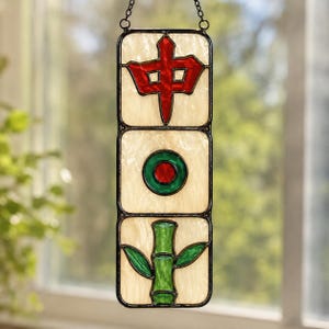 Könnte beinhalten: Ein vertikaler Buntglas-Suncatcher mit drei übereinander angeordneten rechteckigen Feldern auf cremefarbenem, strukturiertem Hintergrund. Das obere Feld zeigt ein rotes chinesisches Schriftzeichen, das mittlere einen grün-roten Kreis und das untere einen grünen Bambusstängel.