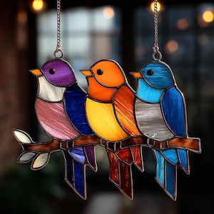 Puede incluir: Obra de arte en vidriera que representa tres pájaros coloridos posados en una rama marrón. Los pájaros son de tonos morados, naranjas y azules, con una variedad de colores en sus alas y colas. La pieza cuelga de una cadena.