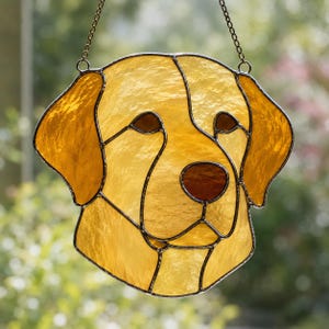 Op de afbeelding: Een glas-in-lood zonnevanger in de vorm van een gele Labrador-kop. Het stuk bestaat uit getextureerde amberkleurige en gouden glassegmenten verbonden door donkere metalen soldeerlijnen, met een donkerbruine neus en ogen, opgehangen aan een dunne metalen ketting.