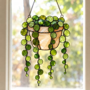Pode incluir: Uma planta suspensa de vitral com um vaso de cor pêssego claro e vinhas pendentes de folhas circulares de vidro verde. A peça está suspensa por uma corrente de metal fina, com cada elemento de vidro emoldurado em solda de metal escuro.