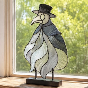 May include: A stained glass plague doctor figure featuring a black top hat, a white beak mask, and a flowing cloak made of textured grey, white, and dark blue glass panels. The piece is mounted on a rectangular black metal base for display.