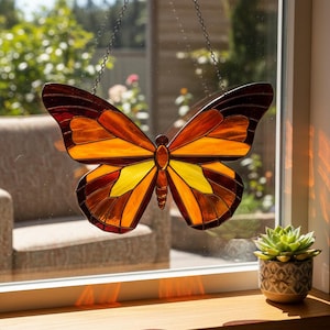 Pode incluir: Apanhador de sol de borboleta em vitral com uma paleta de cores quentes de âmbar, laranja e amarelo. A borboleta é suspensa por uma corrente e pendurada em frente a uma janela. Um detalhe decorativo para a decoração da casa.