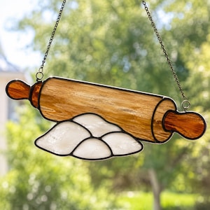 Puede incluir: Un atrapasol de vidrieras con forma de rodillo de madera hecho de vidrio color ámbar con un montón de harina de vidrio blanco debajo. La pieza está enmarcada en soldadura de metal negro y cuelga de una fina cadena de metal negro.