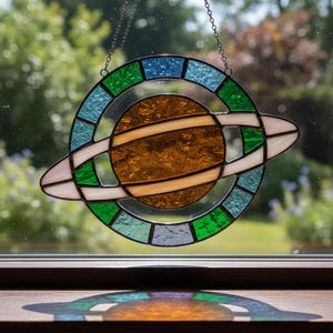 Könnte beinhalten: Glasfenster-Suncatcher mit einem braunen Planeten und einem Ring, umgeben von grünen, blauen und weißen Segmenten. Es hängt an einer Kette. Der Suncatcher befindet sich vor einem Fenster.