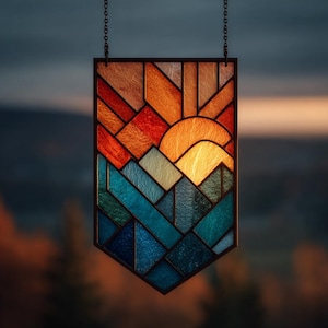 May include: A stained glass panel depicting a sunset over mountains. The design features geometric shapes in shades of orange, red, blue, and green, with a bright yellow sun. The panel is framed in black and hangs from a chain.