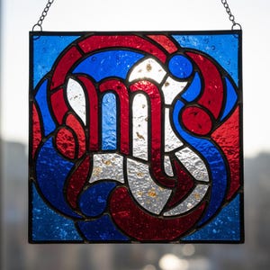 Peut inclure: Panneau carré en vitrail avec une bordure noire, présentant un motif rouge, blanc et bleu. Le motif comprend des lignes et des formes courbes, avec une lettre "M" centrale. Le panneau est suspendu par une chaîne.