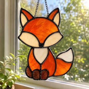May include: A stained glass suncatcher shaped like a sitting fox. The fox features vibrant orange textured glass for the head and body, with cream-colored glass for the face, chest, and tail tip. Dark brown glass forms the paws, all outlined in black.