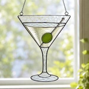 May include: A stained glass suncatcher shaped like a martini glass with a textured clear glass body and a single solid green glass olive. The piece is framed in dark metal solder lines and hangs from a thin metal chain for display in a window.