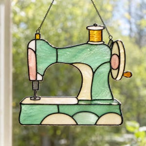 May include: A stained glass suncatcher shaped like a sewing machine, featuring mint green, cream, and soft pink glass panels with a golden yellow spool on top. The piece is outlined in dark metal solder and hangs from a thin black metal chain.