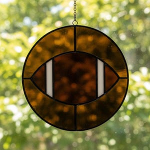 May include: Stained glass football-shaped suncatcher. The design features a brown football shape with white stripes, encased in a circular frame of amber-colored glass. A black chain is attached for hanging. Decorative sports-themed item.