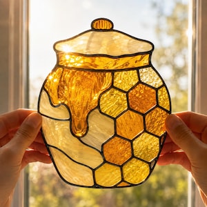 May include: A stained glass suncatcher shaped like a honey pot with a lid. The piece features translucent amber, gold, and clear glass segments arranged in a honeycomb pattern, with a dripping honey design at the top, held against a bright window.