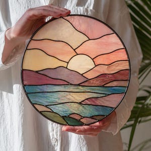 Op de afbeelding: Rond gebrandschilderd glaskunstwerk dat een zonsondergang boven water uitbeeldt. Het kunstwerk heeft een kleurenpalet van roze, oranje, geel, paars en blauw. Het stuk wordt voor een wit overhemd gehouden.