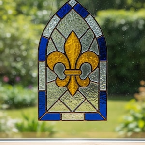 Puede incluir: Panel de ventana de vidrieras con un diseño de flor de lis. El panel tiene un borde arqueado azul y blanco con una flor de lis amarilla en el centro. El vidrio tiene una apariencia texturizada y el panel está suspendido por un alambre.
