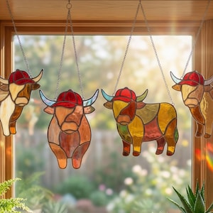 May include: Four stained glass Highland cow ornaments hang from chains in front of a window. Each cow wears a red baseball cap and is made of various shades of brown, yellow, and white glass.