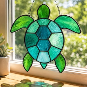 May include: A stained glass turtle suncatcher with a black outline and chain for hanging. The turtle features shades of green and teal glass. The suncatcher is displayed in front of a window, casting a shadow on the wooden surface.