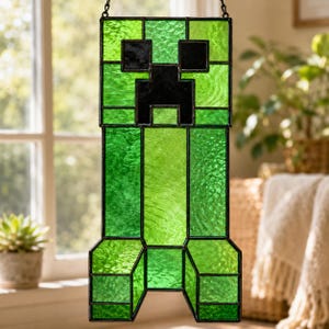May include: A rectangular stained glass suncatcher featuring a green and black pixelated creature design. The piece is constructed from various shades of textured green glass panels joined by dark metal solder lines, forming a blocky character.