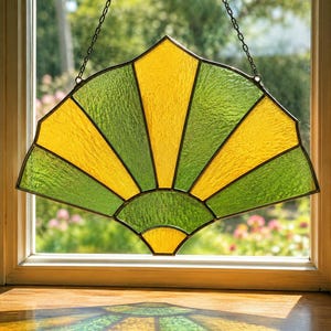 Op de afbeelding: Een gebrandschilderd glazen raam in de vorm van een waaier, met een geel en groen kleurenschema. Het waaierontwerp is verdeeld in afwisselende gele en groene secties, met een donker metalen frame en ketting om op te hangen. Het raam zit in een houten frame.