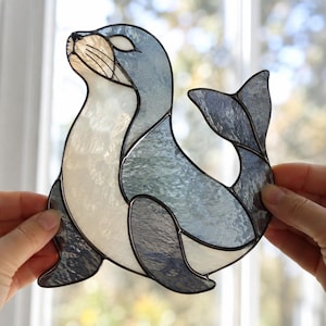 Puede incluir: Un atrapasoles de vidrieras con forma de foca, que presenta piezas de vidrio texturizado en tonos blanco translúcido, azul claro y gris oscuro. Los segmentos de vidrio individuales están unidos por líneas de soldadura de metal oscuro.