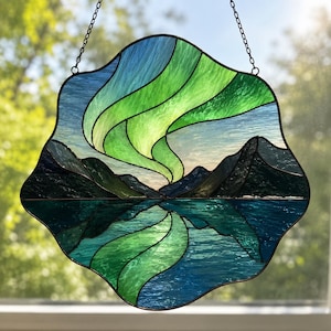 Könnte beinhalten: Ein Buntglas-Sonnenfänger mit einer Landschaft aus dunkelblauen Bergen und einem spiegelnden See unter einem leuchtend grünen und blauen Polarlicht-Himmel. Das Stück hat einen gewellten Rand und ist an einer dünnen Metallkette aufgehängt.