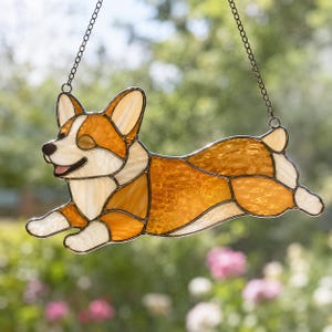 Puede incluir: Un atrapasoles de vidrieras con forma de perro Corgi en posición de carrera. La pieza presenta segmentos de vidrio texturizado en color ámbar, blanco y crema unidos por líneas de soldadura de metal oscuro, suspendida por una fina cadena metálica.