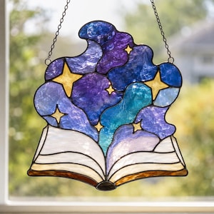 Puede incluir: Un atrapasoles de vidrieras que presenta un libro abierto en la base con páginas blancas. Nubes arremolinadas de vidrio azul profundo, morado y verde azulado se elevan de las páginas, acentuadas por varias estrellas amarillas de cuatro puntas.