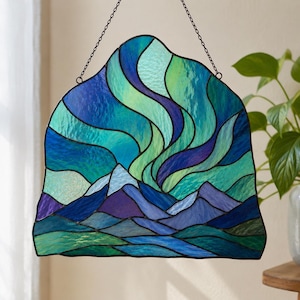 May include: A stained glass suncatcher featuring a landscape of dark blue and purple mountains under a vibrant sky of swirling green, teal, and violet light patterns. The piece is framed in dark metal solder lines and hangs from a thin metal chain.