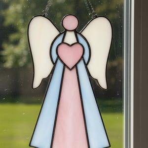 May include: Stained glass angel suncatcher with a heart-shaped center. The angel features white wings, a pink heart, and a light blue and pink dress, all outlined in black. It hangs from a chain.