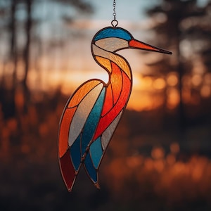 May include: A stained glass bird suncatcher with a long beak and curved neck. The bird features sections of blue, white, orange, red, and yellow glass, outlined in a dark metal frame. It hangs from a chain, with a blurred sunset background.