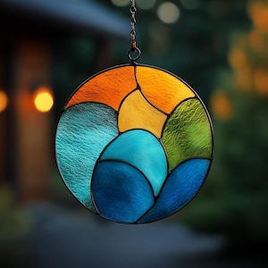 May include: A round stained glass suncatcher with a black metal frame. The design features sections of orange, yellow, green, and blue glass. The suncatcher is suspended by a chain, and the background is blurred.