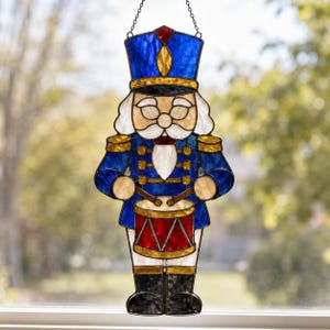 May include: A stained glass nutcracker suncatcher featuring a blue uniform with gold accents, a red drum, and a white beard. The figure wears a tall blue hat with a red top detail and is suspended by a thin metal chain for hanging in a window.