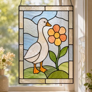 Op de afbeelding: Gebrandschilderd glaspaneel met een witte gans met oranje poten en snavel, naast een grote oranje en gele bloem. De achtergrond toont een blauwe lucht en groene heuvels. Het paneel is omlijst met een zwarte rand.