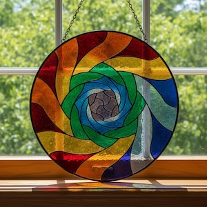 Op de afbeelding: Een rond gebrandschilderd glazen raam met een spiraalvormig ontwerp. Het raam heeft een levendige reeks kleuren, waaronder rood, oranje, geel, groen, blauw en paars. Het glas is omlijst met een zwarte rand en wordt opgehangen door een ketting.