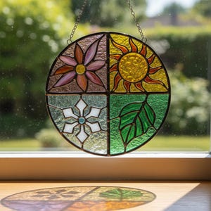 May include: A round stained glass window depicting the four seasons. The top left quadrant shows a pink flower, the top right a yellow sun, the bottom left a white snowflake, and the bottom right a green leaf. The window is hanging in front of a window.
