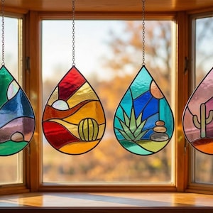 Könnte beinhalten: Vier tropfenförmige Glasfenster-Suncatcher hängen in einem Fenster. Jedes zeigt eine Wüstenlandschaft mit Kakteen, Bergen und Sonnen in Grün, Blau, Rot, Orange und Lila. Sie sind an Ketten aufgehängt.