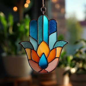 May include: Stained glass Hamsa hand suncatcher with a copper-colored frame. The hand features a colorful design with shades of blue, teal, orange, yellow, and pink. The suncatcher is suspended by a thin, gold-colored chain.