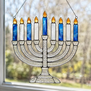 May include: A stained glass menorah suncatcher featuring nine candles with amber flames. The candles alternate between blue and white glass, while the base and arms are constructed from textured clear glass with dark metal solder lines.