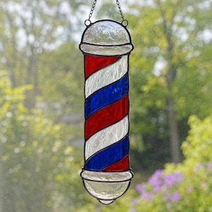Peut inclure: Un ornement de poteau de barbier en vitrail avec des rayures diagonales en verre rouge, blanc et bleu. La pièce cylindrique possède un dôme en verre texturé transparent en haut et en bas, assemblé par des lignes de soudure en métal sombre.