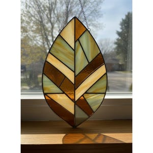 May include: Stained glass leaf with a geometric design. The leaf features a mix of amber, light green, and cream-colored glass pieces, outlined in black. A silver chain is attached for hanging. The leaf is approximately 10 inches tall.