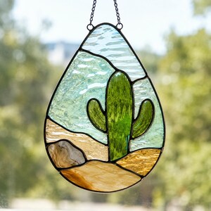 May include: A teardrop-shaped stained glass suncatcher featuring a central green saguaro cactus. The background consists of textured light blue glass representing the sky, while the foreground features tan and brown glass depicting desert terrain.