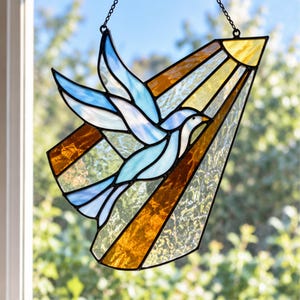 Puede incluir: Un atrapasueños de vitral con un pájaro azul claro en vuelo sobre un fondo de rayos de vidrio texturizado ámbar y transparente. La pieza está enmarcada con líneas de soldadura de metal negro e incluye una pequeña cadena metálica para colgar.
