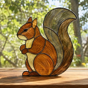 Könnte beinhalten: Eine Eichhörnchenfigur aus Buntglas, gefertigt aus strukturierten bernsteinfarbenen, braunen und cremefarbenen Glasstücken, die durch dunkle Metalllötlinien verbunden sind. Das Eichhörnchen ist im Profil sitzend mit einem großen, buschigen Schwanz dargestellt.